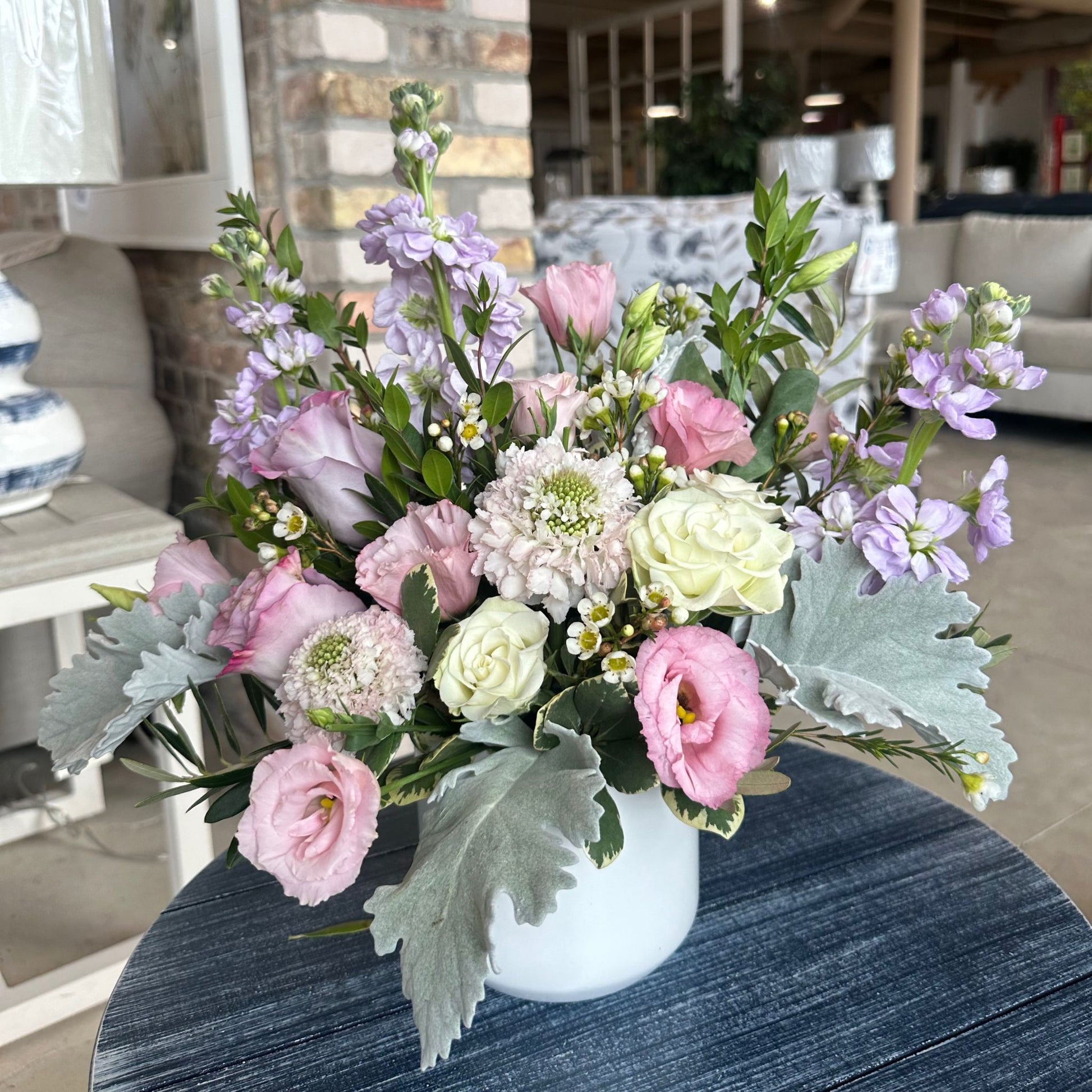 Ever Enchanted floral arrangement by Birch & Blossom with lavender roses, pink scabiosa, lavender stock, white spray roses, pink lisianthus, wax flower, dusty miller, pittosporum, and myrtle in a white ceramic container. Soft and romantic hues.
