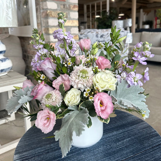 Ever Enchanted floral arrangement by Birch & Blossom with lavender roses, pink scabiosa, lavender stock, white spray roses, pink lisianthus, wax flower, dusty miller, pittosporum, and myrtle in a white ceramic container. Soft and romantic hues.