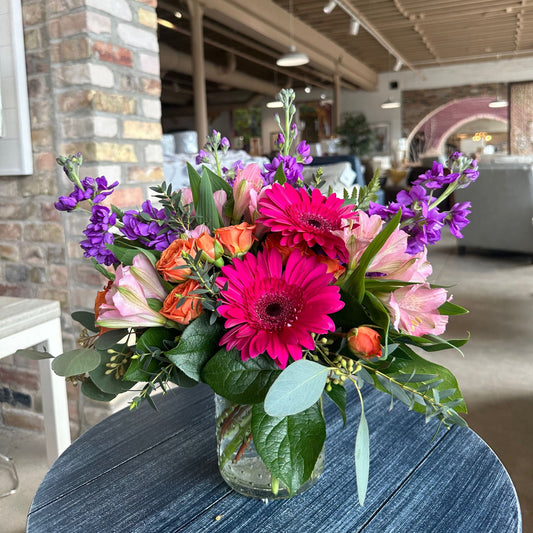 Girly POP! energetic floral arrangement by Birch & Blossom with fuchsia gerbera daisies, orange rose spray, pink alstroemeria, purple stock, salal, eucalyptus, and variegated pittosporum in a vase. Fun and vibrant colors.